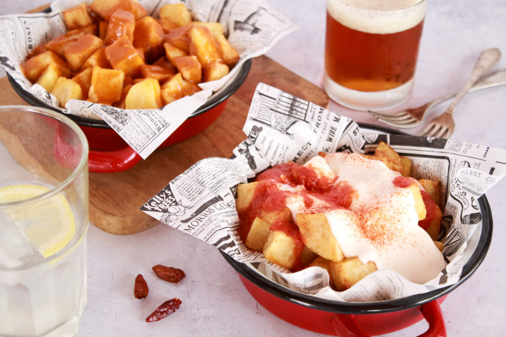 Dos raciones de patatas fritas en dados grandes servidas una con salsa brava y la otra con veganesa y salsa picante de tomate