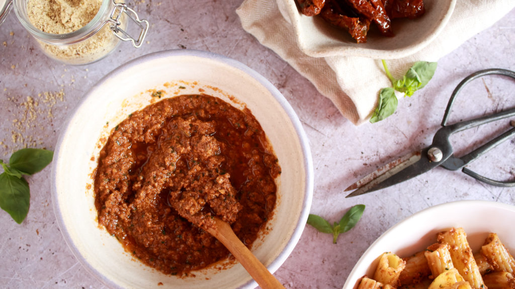 Pesto rosso en un bol con una cuchara de madera y alrededor tomates secos en aceite, pasta y pasrmesano vegano