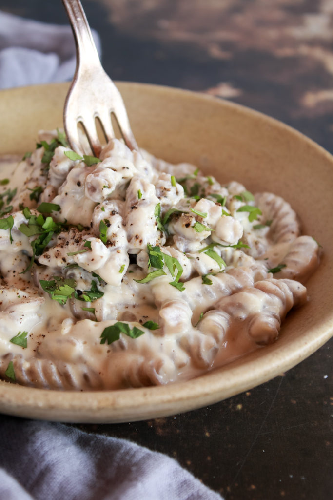 Un plato de pasta fussilli de trigo sarraceno con salsa Alfredo vegana. Un tenedor pincha la pasta.