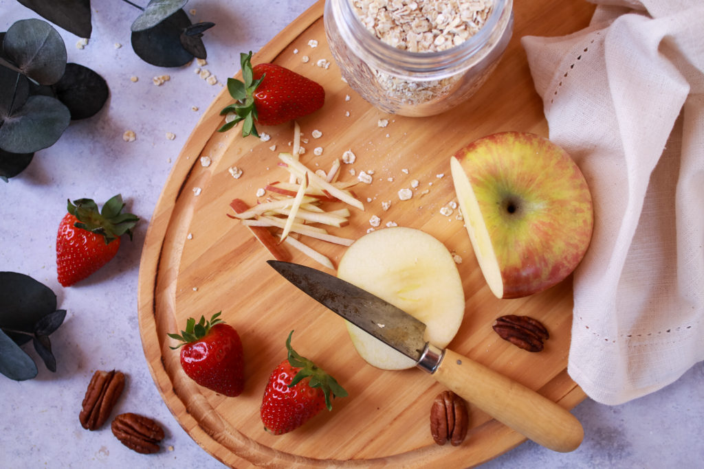 Bodegón de fruta con manzana cortada, fresas y almendras junto a un bote con copos de avena suaves