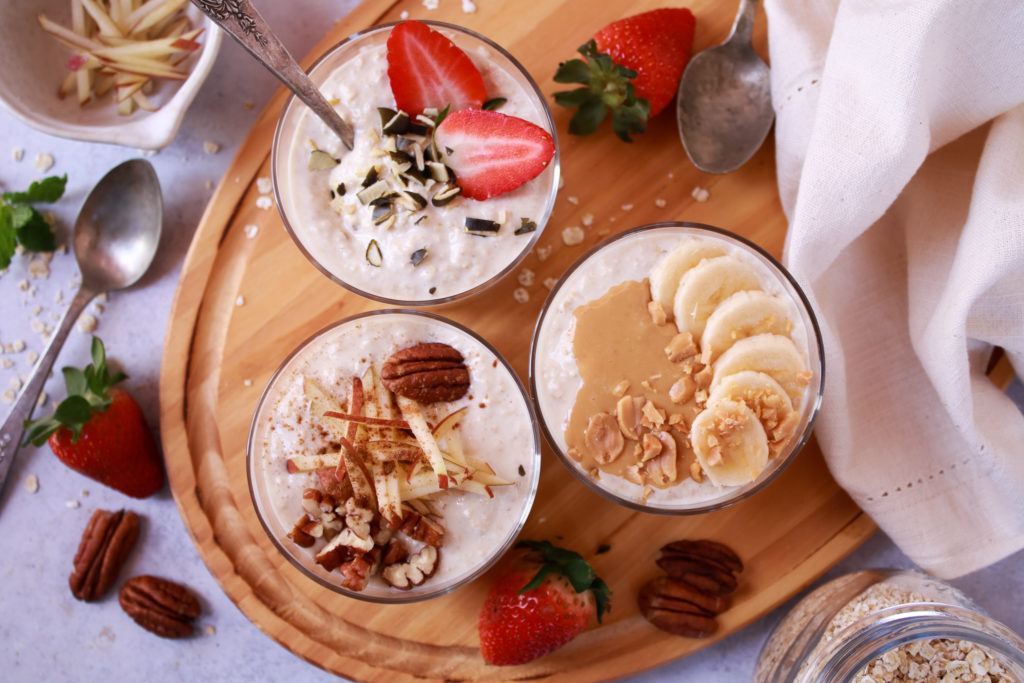 Foto horizontal con tres vasos con muesli bircher y con diferentes toppings, uno con cacahuete y plátano, otro con fresas y semillas de calabaza y un último con manzana en bastones, nueces pecanas y canela