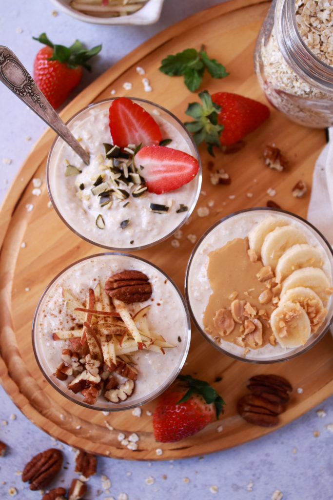 Foto vertical con tres vasos con muesli bircher y con diferentes toppings, uno con cacahuete y plátano, otro con fresas y semillas de calabaza y un último con manzana en bastones, nueces pecanas y canela